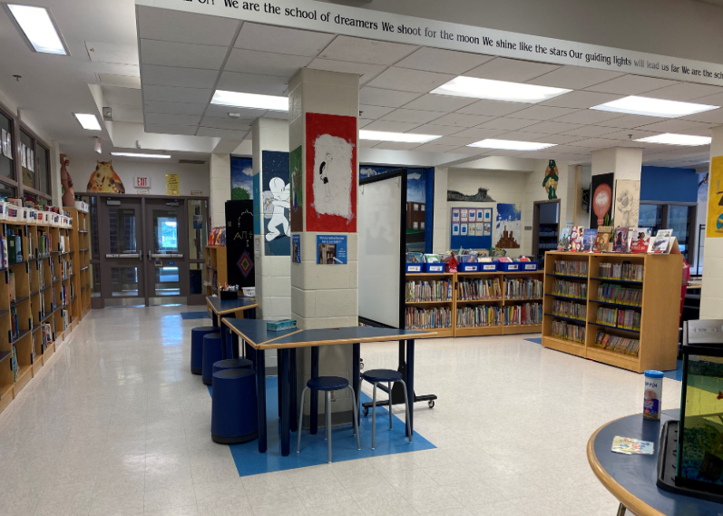 shelves of books line the wall. A table with stools is in the centre of the room with pillars featuring posters. more bookshelves can be seen on the other side.